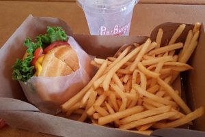 Eine Kartonschale in welcher sich Pommes und ein Burger befinden. Der Burger ist mit Salatblättern, Tomaten und Käse belegt und ist in einer Papiertüte. Im Hintergrund ist ein Trinkbecher aus Plastik zu sehen.
