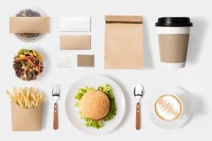 Ein Salat in einer Schale, Pommes in einer Papiertüte, ein Teller mit einem Burger, daneben Gabel und Löffel. Eine Tasse mit Kaffee, ein Kaffee to go Becher, eine Papiertüte und weitere Verpackungen aus Papier.