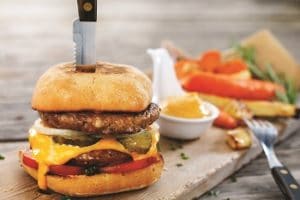 Ein vegetarischer Burger von Quorn, belegt mit einer vegetarischen Bulette, Käse, Tomaten und Gurken. Der Burger ist mit einem Messer aufgespießt.