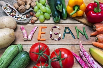 Auf dem Foto ist mit verschiedenen Gemüsesorten das Wort "vegan" auf einem Holzfußboden ausgelegt. Um den Schriftzug herum befindet sich Obst und Gemüse, wie zum Beispiel Tomaten und Zucchini in der unteren Bildhälfte und Paprika, Zucchini, Nüsse und Weintrauben in der oberen Hälfte des Bildes.