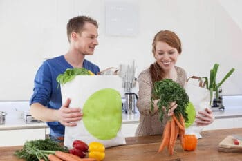 Zwei Menschen stehen in der Küche mit einer Papiereinkaufstüte und viel Obst / snackconnection