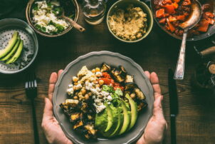 Female hands holding with healthy vegetarian bowl with various grilled vegetables, avocado and chickpea hummus on rustic background, top view. Clean food and dieting nutrition concept / snackconnection