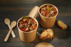 Two unlabelled takeaway tubs of rich beef goulash with spicy chili peppers served with toasted baguette and plastic spoons on a rustic wooden table with copyspace viewed high angle
