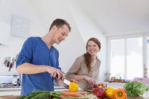 Zwei Menschen stehen in der Küche und lachen beim Gemüse schneiden / snackconnection